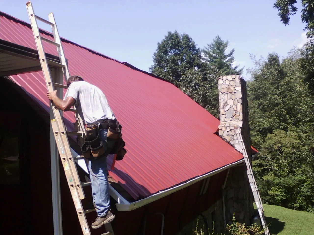 Roofer on a ladder beside a red metal roof and stone chimney — Soffit & Fascia Repair by Roofing Brothers in Dorr